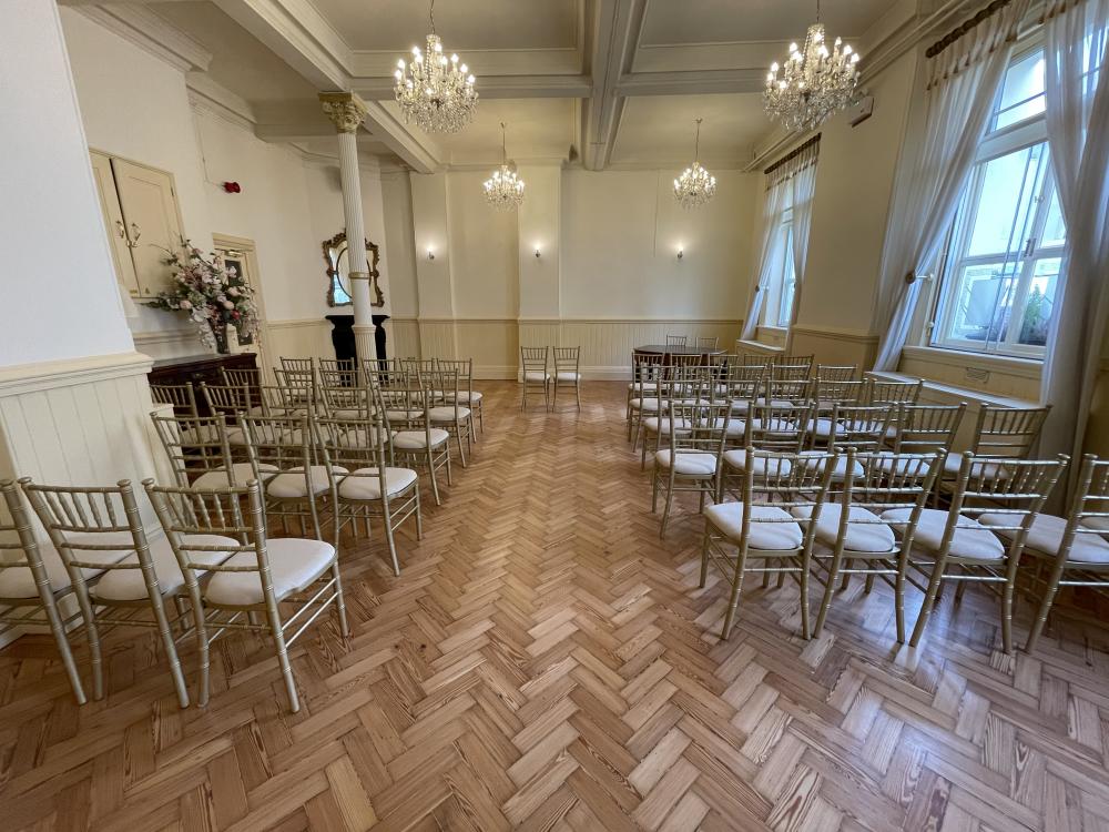 The Regency Room. Two rows of chairs with an aisle in the middle. Chandeliers on the ceiling. Open windows