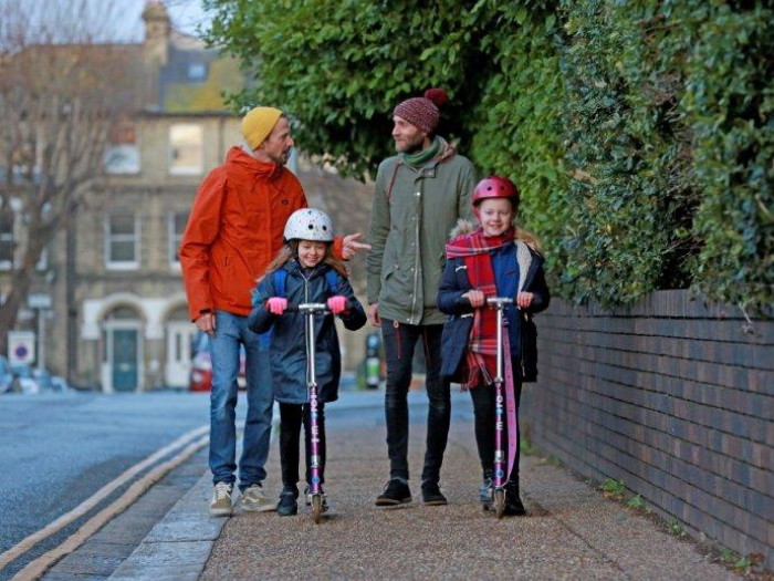 "Two men walking on a pavement behind two schoolgirls on scooters"