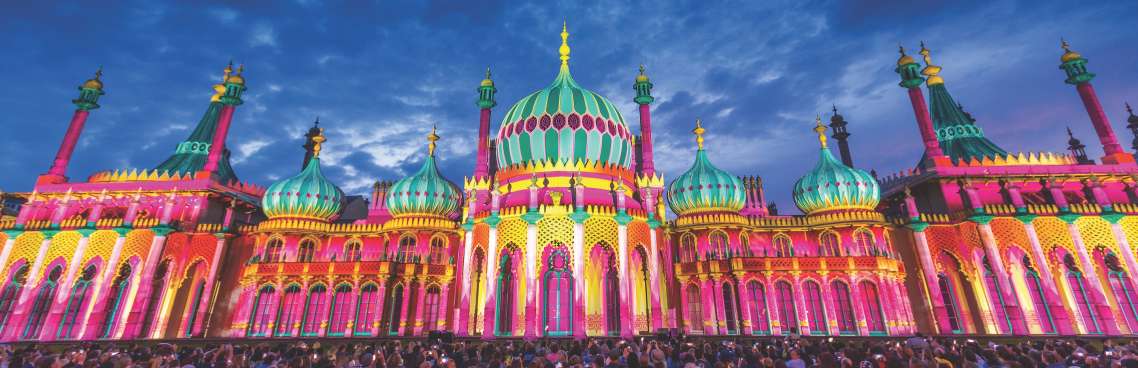 Brighton Pavillion lit with multicoloured lights at night. A crowd of people are admiring and taking photos of the building lit up.