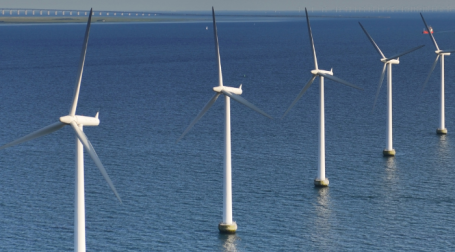 Five tall, white, 3-blade wind turbines protrude from the sea. They stand in a line stretching off into the distance.