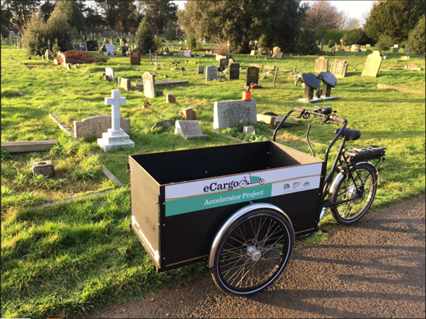 An eCargo bike is in a cemetery. The bike consists of a normal front wheel and pedals, then the frame becomes a cart, suitable for transporting goods, with a wheel either side.