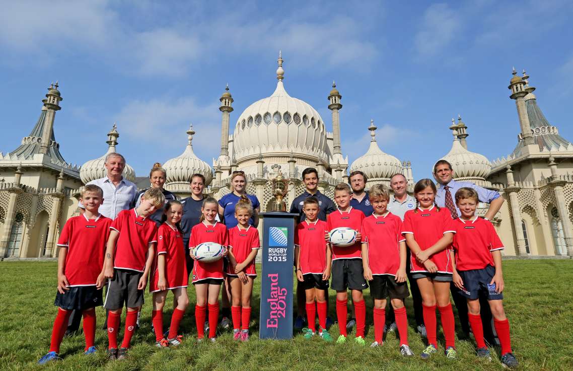 ten children dressed in a red rugby kit line up for a photo with the rugby world cup. Eight adults stand behind them, some dressed for sport, others in shirts and ties. Two childen hold rugby balls, and in the centre is a blue podium with the golden cup on top of it. In the background is the Brighton Pavilion with its distinctive Dome.