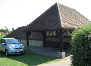 A tiled roofed shelter, open on three sides. Large wooden pillars at each corner support the eaves. A wooden bench runs along the length of the back wall.