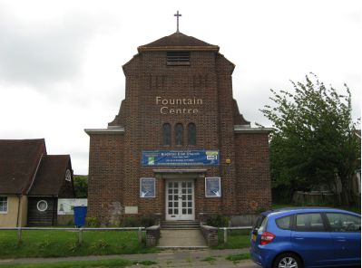 A brickwork church front, with small arched windows and a cross at the top. The entrance is white double doors. The name 'Fountain Centre' is written on the front in gold