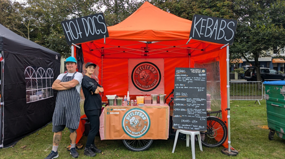 Food cart with chalkboard menu next to it. Everything is under an orange gazebo. Two people stand back to back with their arms crossed.