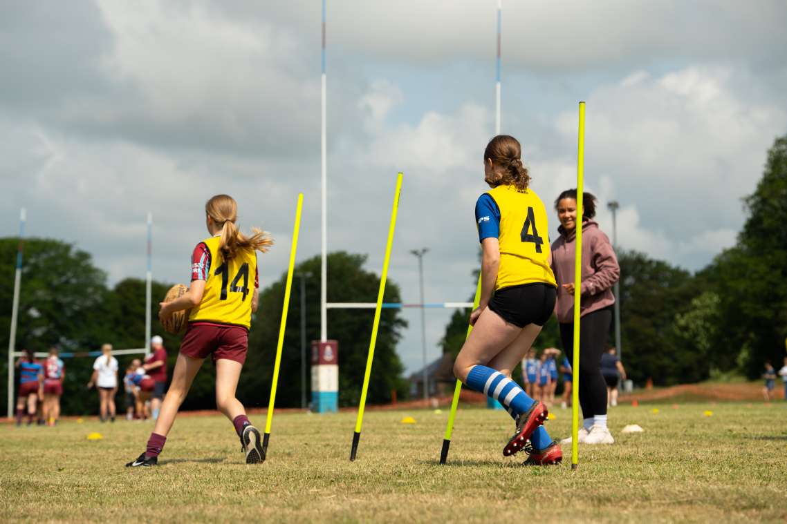 Two girls - one carrying a rugby ball and both wearing yellow bibs - weave through a series of yellow poles while being encouraged by a women looking on