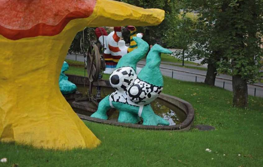 Pale green humanoid sculpture doing a handstand in a black and white patterned bathing suit