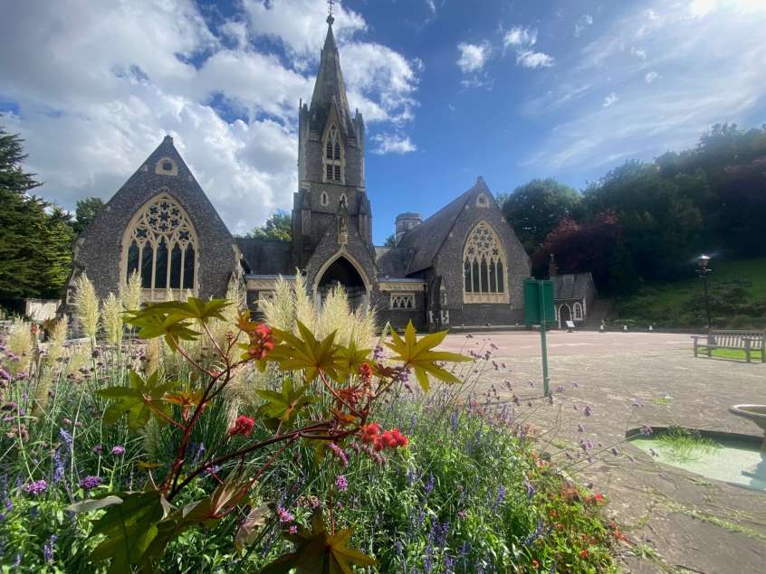 Main entrance of Woodvale Crematorium. Flowers in the foreground, and a large stone building with intricate windows in background.