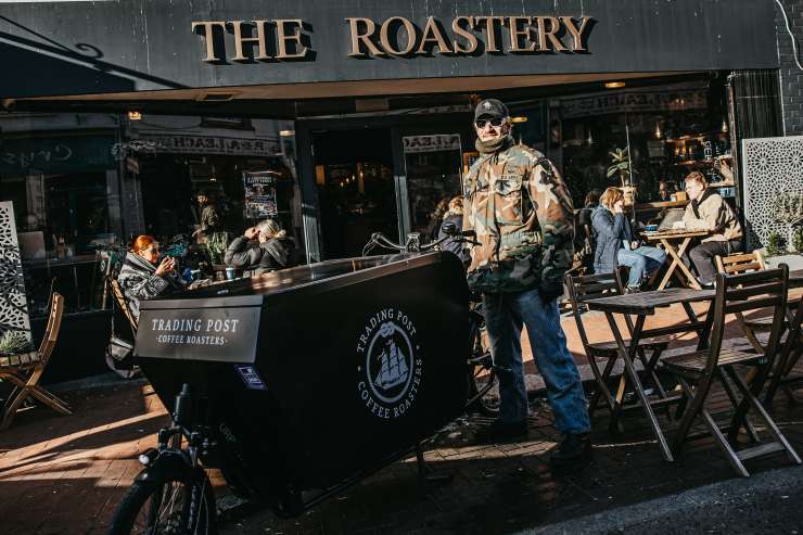 A man smiles as he stands behind an ecargo bike branded with the Tranding Post Coffee Company logo.