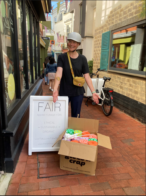 A person wearing a helmet stands in a redbrick shopping alley. Behind them is an ebike. There is a box of sustainable toilet paper at their feet.