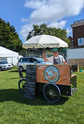 Beelzebab cart. Chalkboard with menu in front. Man standing behind the cart. Sun umbrella shading him. 