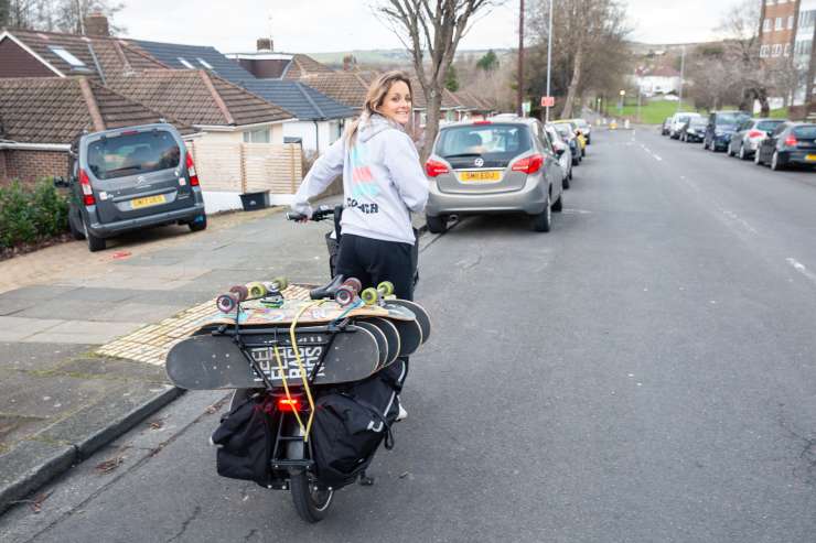 A rear shot of Sarah on her ecargo bike looging behind her into the camera, she is smiling. There are a number of colourful skateboards straped to the back of the bike.