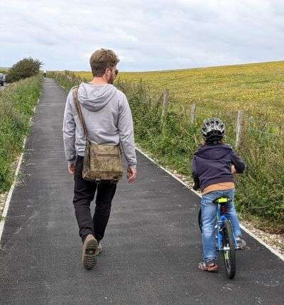 The rear view of a man and walking next to a small child on a bike. They're on a footpath in the Brighton countryside. 