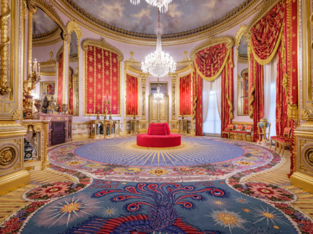 The recently restored Saloon at the Royal Pavilion, showing the intricately patterned carpet, scarlet and gold curtains and chandelier hanging from a ceiling painted with a blue sky and clouds.