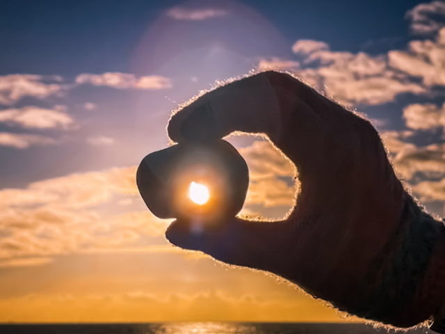 A hand holding a pebble up to the sunset at the beach. Photo credit @BenJamesPhotos