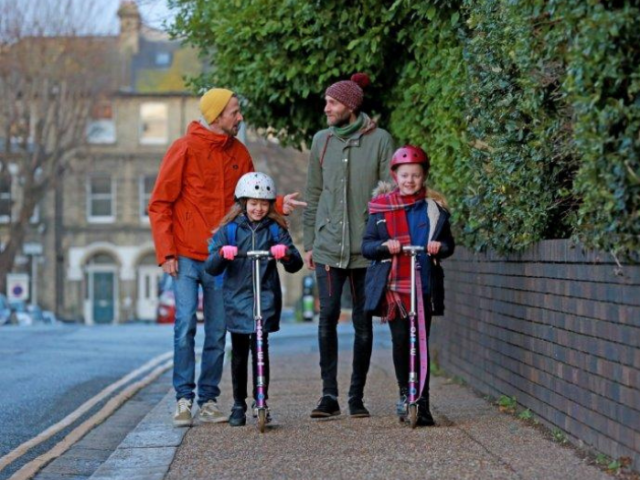 Two fathers walking on a pavement with their daughters on scooters in front of them