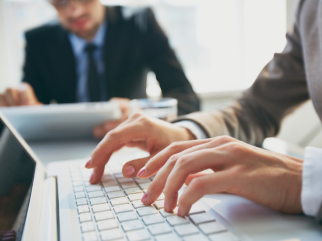 Person's hands typing on a computer keyboard, with man in a suit in background sitting in front of another laptop.