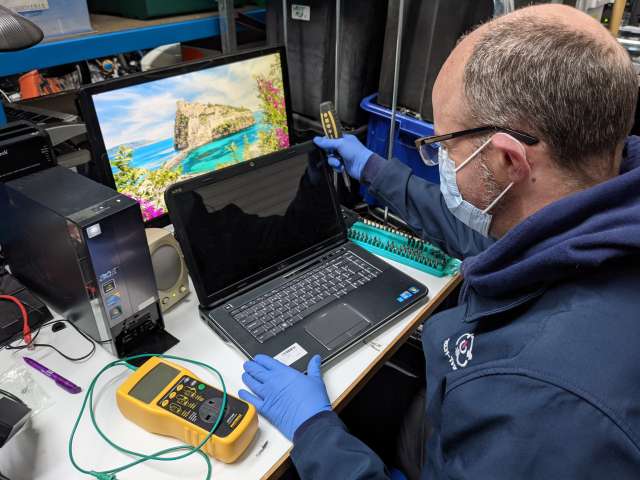 Man wearing gloves and masks seated in front of a laptop with another behind in a repair workshop. 