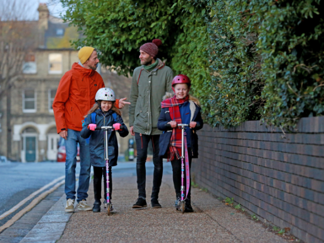 A picture of two men walking with two children on scooters in front of them