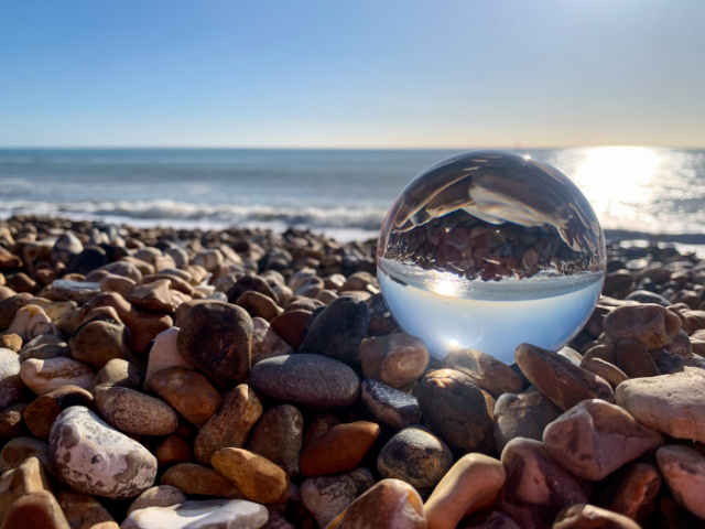 Pebbles on seashore, with sea in the background and a glass globe half-filled with water in the foreground