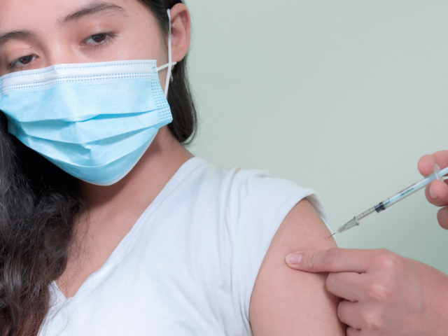 A masked teenage girl on the left has her vaccination in her upper left arm from an unseen person to the right 