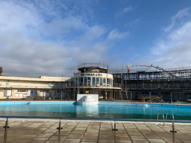 In the foreground are grey floor tiles in front of the open air lido swimming pool. The pool has a light blue wall enclosing it and white steps leading into it directly opposite. Behind this is a semi-circular two story building in art deco style, with the sign "Saltdean Lido" abouve the tall windows. The building is surrounded by scafolding.