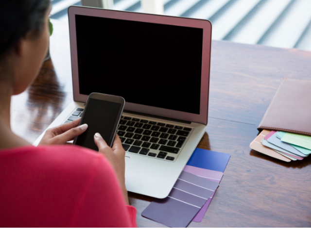 Woman in pink top scrolling on a mobile phone with laptop and different coloured cards in the background