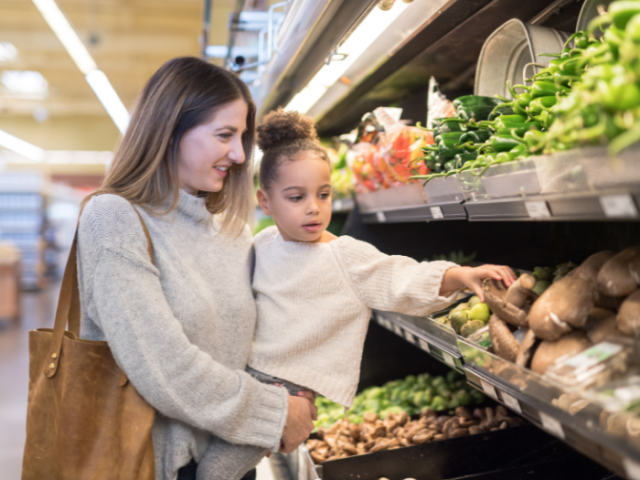A young woman carries a young girl while shopping in a supermarket. The girl points out fruit and veg for sale. 