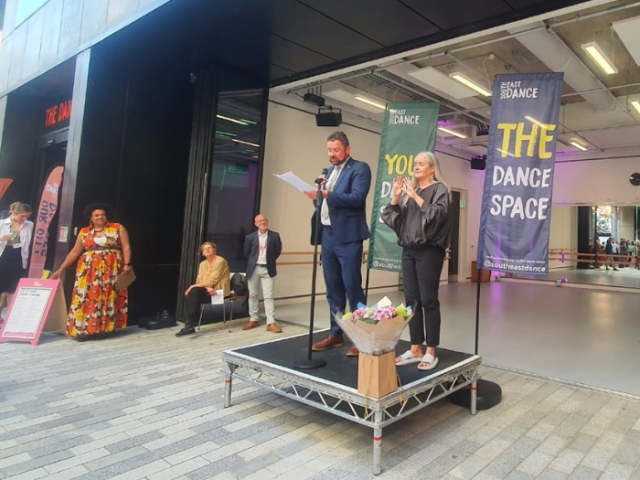 A man and a women are stood on a low, square black platform in front of a large glass fronted building. The building has banners up in it with the wording "The Dance Space". On the stage Cllr Phelim Mac Cafferty is giving a speech and has notes in his hand. he is standing at a microphone.Beside him is a sign-language expert and on the stge in front of them is a bunch of flower.