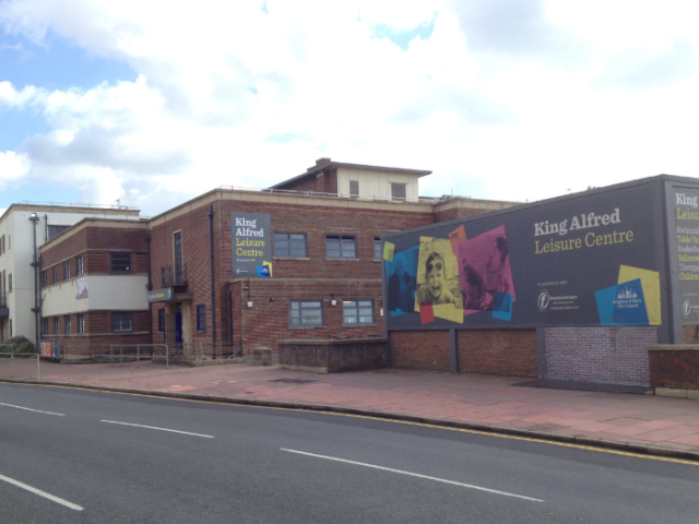 The King Alfred Leisure Centre pictured from the other side of the road.The brick built building is in for stages with a flat roof. A white clad building at the end on the left, is next to another with a white rendered band between its two floors. Then comes an extension featuring the entrance below a grey canopy. Finally, on the left a lower building is faced with a grey sign features 3 large colour images of people enjoying the centre's activities.