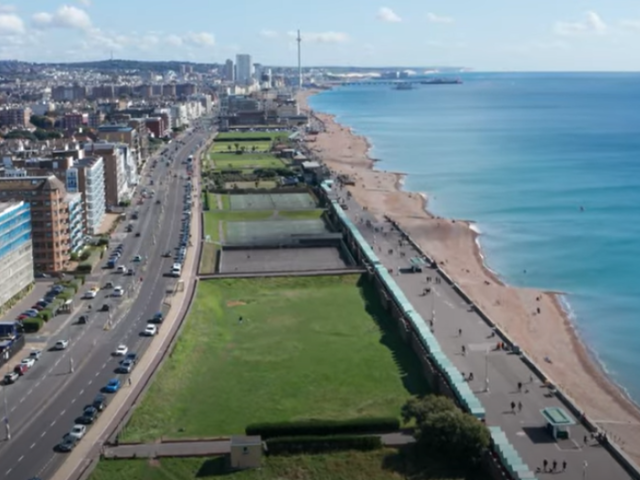 The picture is an aerial view of Hove Lawns stretching east towards central Brighton. In the cnetre of the image is the continuous green strip of lawns punctuated at inttervals by three tarmac areas. To the right is the beach and the sea. To the left vehicles drive up and dwon the coastal raod, which on its left is framed by blocks of flats in shades of white, brown and blue. In the distance the tower of the i360 is just visible.