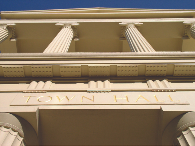 A picture of the front elevation of Brighton Town Hall showing two pairs of white columns supporting the entrance arch and the roof of the building. The words "Town Hall" are inscribed between the lower pair