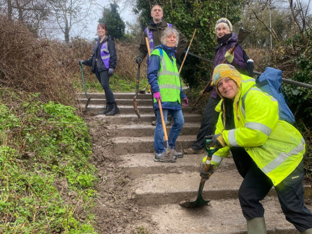 Photograph of a group of volunteers wearing hi vis jackets on some muddy steps. They are all using tools to remove weeds from the steps.