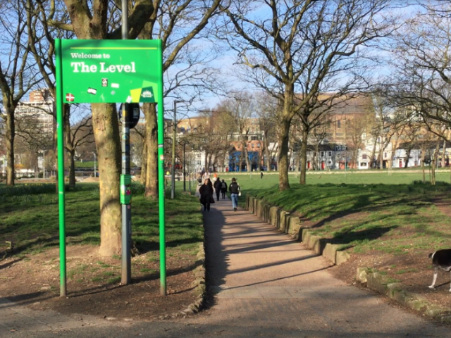 Photograph of a green tall welcome sign and a brick path leading to a large green space with trees.