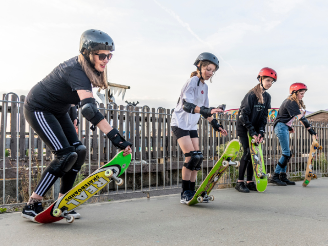 Picture shows four young women wearing helmets and protective gear, getting ready to ride a skateboard.