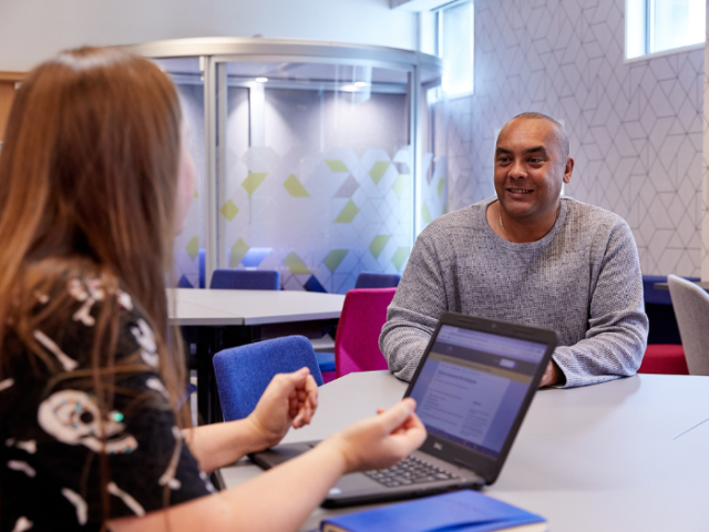 The photograph shows a woman with her laptop open, giving advice to a smiling man.