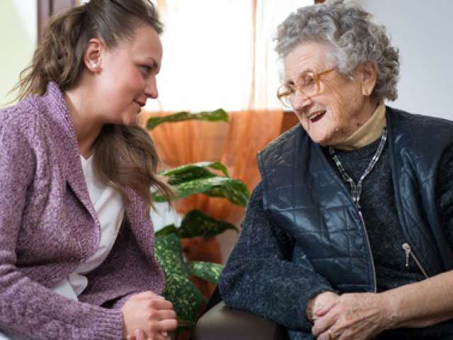 Image of care worker and elderly woman sitting next to each other and laughing