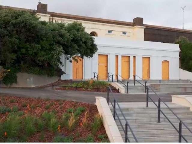 The renovated reading room appears as a white fronted building at the lower level with concrete steps leading up towards five tall window/doorways still currently closed with yellow baording. The second level is just visible recessed back and bordered by trees