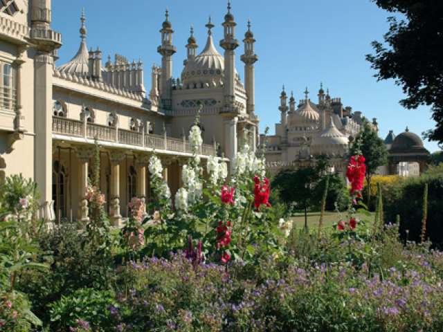 In the foreground a border of red and purple flowering plants are enjoying a sunny day. Behand the planting the domes of the Royal pavillion can been seen against a blue sky.