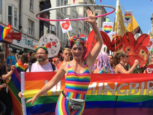 Person in rainbow outfit with a hula hoop in the air and a crowd or people behind them