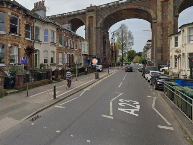 A picture of a road and a viaduct with pavement on either side as well as cars and bins