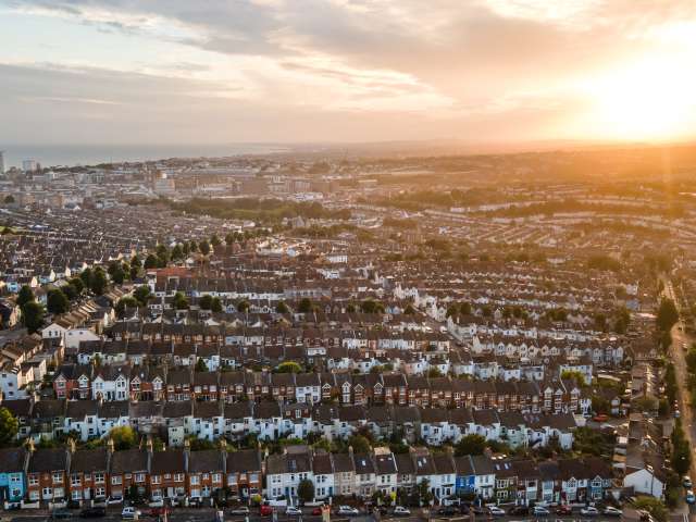 An aerial view of Brighton. Hanover, with its colourful terraced houses, is in the foreground. The view stretches across the city to the sea. The sun is setting behind the downs to the west. The sky is still blue, but the pink hues around the sun suggest a spectacular sunset is coming.