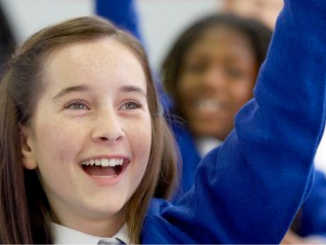 The image shows two young secondary age schoolchildren raising their hands in class.