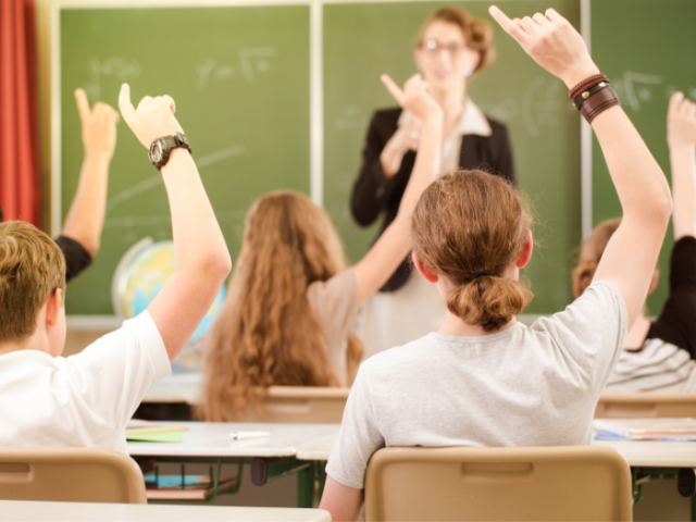 The image shows secondary school pupils raising their hands in class to answer a question.