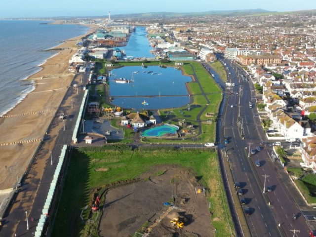 An aerial photograph shows building work taking place on a grassed area in the foreground, with Hove Lagoon and then Shoreham Harbour stretching out into the distance beyond that on a sunny day. The Esplanade path and beach can be seen to the left of the works area and lagoon. 