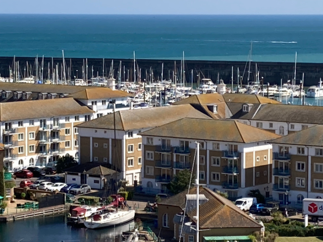 In the foreground two white yatches with burgundy sail covers and awnings are moored in front of a four-story block of apartments. The building has white rendered top and bottom floors, with yellow bring second and thrid floors. Similar designed blocks stretch away from it in paralell and at right angles leads towards the main marina behind. This if packed with moored yatchs, their masts stretchng above the dark grey sea wall beind them. In the distance is the sea. 