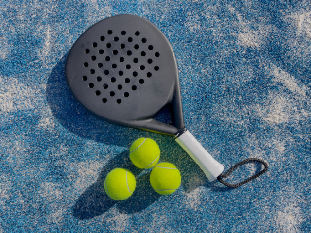 A black and grey padel tennis racquet lays on a mottled blue and white cloth. Beside it our 3 luuminous yellow balls. The racquet head is oval shaped and has holes in a square regular patter across the face. Beneath the white handle is a black chord formed into a loop