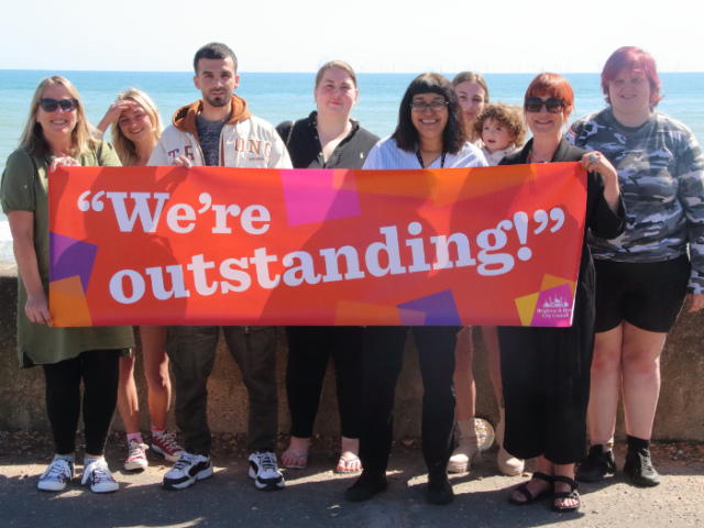 A group of you care leavers and their personal advisors on Brighton & Hove seafront holding a colourful banner reading 'We're outstanding!'