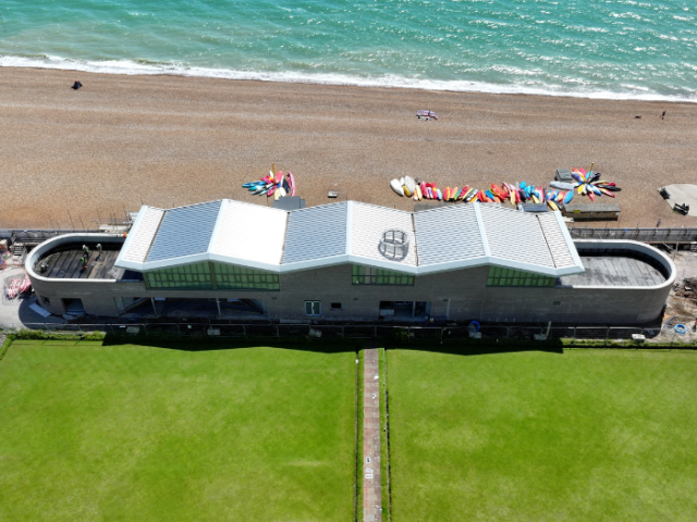 An aerial image of the new Outdoor Sports Hub being built in Hove Beach Park. The silver roof resembles 3 letter w's and there is a semi-citrcular balcony on the upper floor of the 2 storey building at either end. In front of the building the lawns are bisected by a footpath leading to the centre of the building. Behind it is the beach and sea.