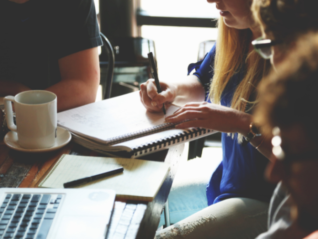 A group of people sat at a table with a laptop, notepads and a mug